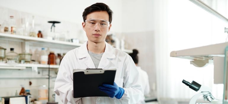 Researcher in a laboratory holding a clipboard while standing in front of laboratory equipment.